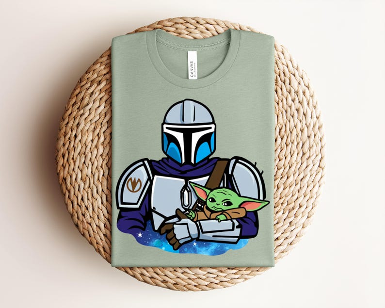 Puede incluir: Camiseta verde salvia con una ilustraci&oacute;n de dibujos animados de un personaje mandaloriano sosteniendo a un alien&iacute;gena verde. El mandaloriano lleva un casco y una armadura plateados con detalles azules. El alien&iacute;gena est&aacute; envuelto en una capa marr&oacute;n.