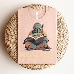 May include: A light peach-colored t-shirt featuring a graphic of a helmeted figure in armor sitting cross-legged and holding an open book with a small green creature. The text "READING IS THE WAY" is printed in a bold, light-colored font below.