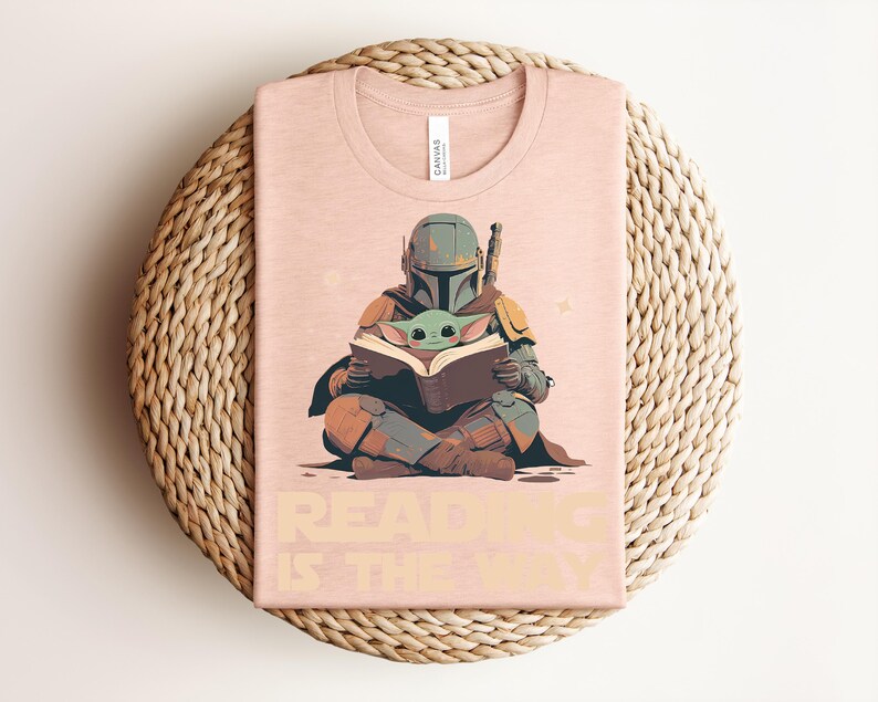 May include: A light peach-colored t-shirt featuring a graphic of a helmeted figure in armor sitting cross-legged and holding an open book with a small green creature. The text "READING IS THE WAY" is printed in a bold, light-colored font below.