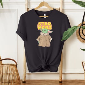 Puede incluir: Camiseta negra con un gr&aacute;fico de Grogu de Star Wars, y el texto "STAR WARS" en amarillo y rosa. Grogu lleva un abrigo marr&oacute;n y tiene la piel verde. La camiseta est&aacute; colgada en una percha de madera.