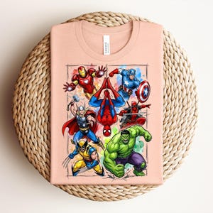 May include: A peach-coloured t-shirt displaying a vibrant graphic of Marvel superheroes, such as Iron Man, Captain America, Spider-Man, Thor, Wolverine, Hulk, and Deadpool. The shirt is placed on a woven, circular surface.