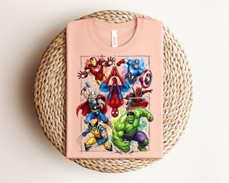 May include: A peach-coloured t-shirt displaying a vibrant graphic of Marvel superheroes, such as Iron Man, Captain America, Spider-Man, Thor, Wolverine, Hulk, and Deadpool. The shirt is placed on a woven, circular surface.