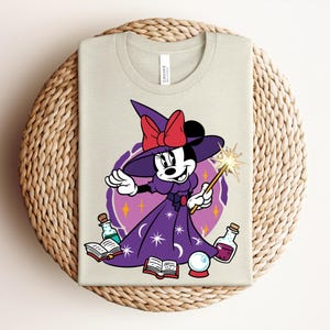 May include: A light beige t-shirt featuring a cartoon Minnie Mouse dressed as a witch. She wears a purple dress and hat with a red bow, holding a wand. The design includes potion bottles, books, and a crystal ball.