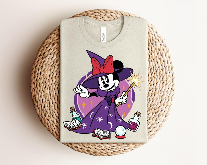 May include: A light beige t-shirt featuring a cartoon Minnie Mouse dressed as a witch. She wears a purple dress and hat with a red bow, holding a wand. The design includes potion bottles, books, and a crystal ball.