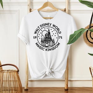 May include: White t-shirt featuring a black graphic of the Walt Disney World Magic Kingdom logo. The design includes a castle, moon, stars, and the years 1971 and the words "Walt Disney World" and "Magic Kingdom".