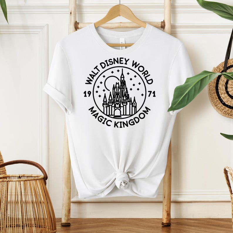 May include: White t-shirt featuring a black graphic of the Walt Disney World Magic Kingdom logo. The design includes a castle, moon, stars, and the years 1971 and the words "Walt Disney World" and "Magic Kingdom".