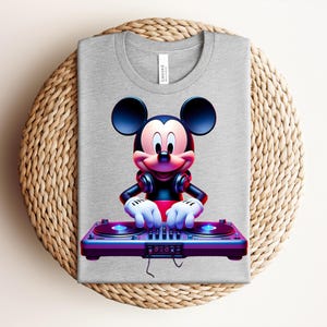 May include: A gray t-shirt featuring a vibrant graphic of Mickey Mouse as a DJ, wearing headphones and operating a turntable. The design is colorful with shades of red, blue, and purple. The shirt is folded on a woven, circular surface.