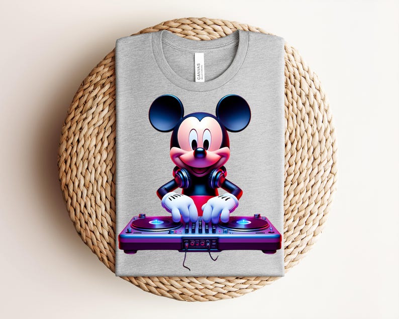 May include: A gray t-shirt featuring a vibrant graphic of Mickey Mouse as a DJ, wearing headphones and operating a turntable. The design is colorful with shades of red, blue, and purple. The shirt is folded on a woven, circular surface.
