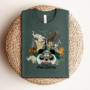 Puede incluir: Una camiseta de cuello redondo de color verde oscuro con un estampado gr&aacute;fico de Mickey Mouse mirando a trav&eacute;s de binoculares rodeado de varios animales, incluyendo un elefante, una jirafa, un tigre y una cebra, con el texto Disney's Animal Kingdom.