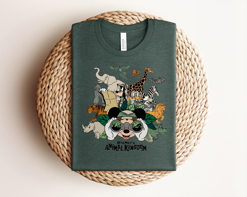 Puede incluir: Una camiseta de cuello redondo de color verde oscuro con un estampado gr&aacute;fico de Mickey Mouse mirando a trav&eacute;s de binoculares rodeado de varios animales, incluyendo un elefante, una jirafa, un tigre y una cebra, con el texto Disney's Animal Kingdom.