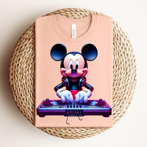 May include: A peach-colored t-shirt featuring a vibrant graphic of Mickey Mouse as a DJ. Mickey is wearing headphones and has his hands on a DJ mixer. The shirt is folded on a woven, circular surface.