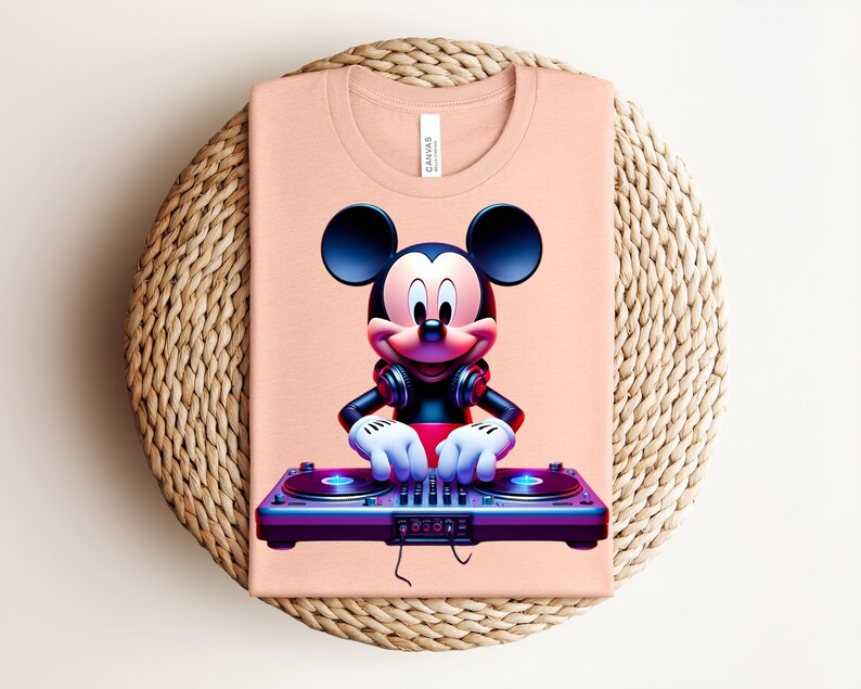 May include: A peach-colored t-shirt featuring a vibrant graphic of Mickey Mouse as a DJ. Mickey is wearing headphones and has his hands on a DJ mixer. The shirt is folded on a woven, circular surface.