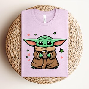 May include: A light purple t-shirt featuring a cartoon character with large green ears, holding a brown cup. The character is wearing a brown coat and a tan scarf. Green stars and pink hearts are scattered around the character.