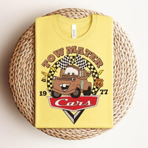 May include: A light yellow t-shirt featuring a graphic of the character Tow Mater from the movie Cars. The design includes the text Tow Mater, the number 1977, a Route 66 sign, checkered flags, a trophy, and the red Cars logo at the bottom.