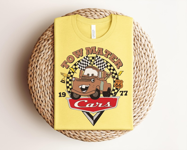 May include: A light yellow t-shirt featuring a graphic of the character Tow Mater from the movie Cars. The design includes the text Tow Mater, the number 1977, a Route 66 sign, checkered flags, a trophy, and the red Cars logo at the bottom.