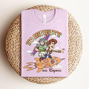 May include: A light purple t-shirt featuring a graphic print of Buzz Lightyear and Woody riding a rocket. The text reads TO INFINITY AND BEYOND in a retro font. The design includes clouds, stars, and orange flames coming from the rocket ship.