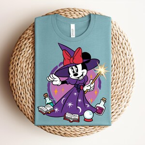 May include: A light blue t-shirt featuring a cartoon Minnie Mouse dressed as a witch. Minnie wears a purple dress and hat with a red bow, holding a wand. The design includes potion bottles, a crystal ball, and open books.