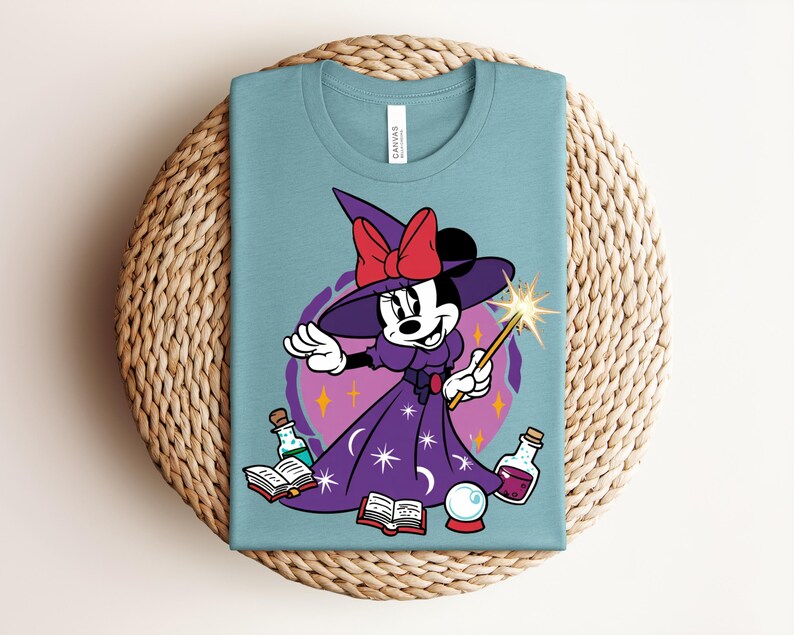 May include: A light blue t-shirt featuring a cartoon Minnie Mouse dressed as a witch. Minnie wears a purple dress and hat with a red bow, holding a wand. The design includes potion bottles, a crystal ball, and open books.
