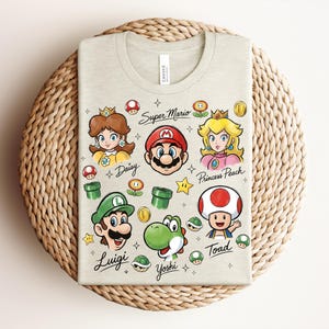 May include: A light beige t-shirt featuring colorful illustrations of Super Mario characters, including Mario, Luigi, Princess Peach, and Yoshi. The words "Super Mario" and character names are written on the shirt. The shirt is folded on a woven, circular surface.