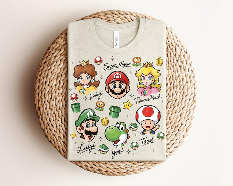 May include: A light beige t-shirt featuring colorful illustrations of Super Mario characters, including Mario, Luigi, Princess Peach, and Yoshi. The words "Super Mario" and character names are written on the shirt. The shirt is folded on a woven, circular surface.
