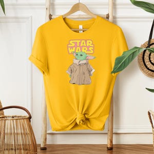 Puede incluir: Una camiseta amarilla con un gr&aacute;fico de Grogu de Star Wars. El dise&ntilde;o incluye el texto "STAR WARS" en una fuente rosa y amarilla encima del personaje. Grogu est&aacute; representado con un abrigo marr&oacute;n y piel verde.