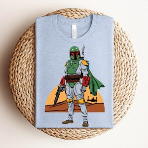 May include: Light blue t-shirt featuring a cartoon illustration of a character in green and gray armor, holding a weapon. The character stands in front of a desert landscape with a setting sun and a spaceship. The shirt is folded on a woven surface.