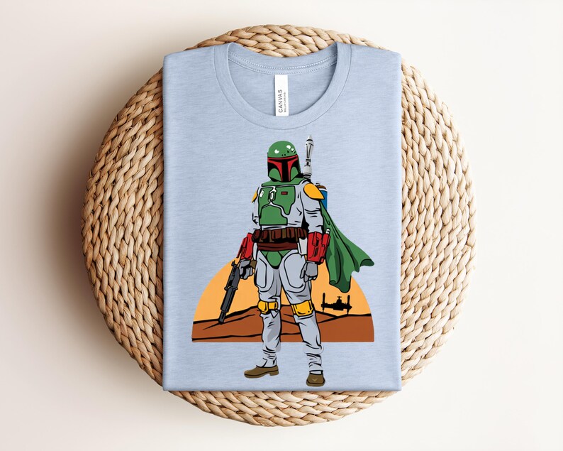 May include: Light blue t-shirt featuring a cartoon illustration of a character in green and gray armor, holding a weapon. The character stands in front of a desert landscape with a setting sun and a spaceship. The shirt is folded on a woven surface.