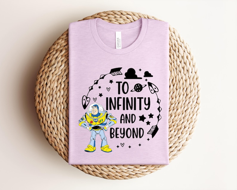 Puede incluir: Una camiseta de color morado claro con un gr&aacute;fico de un personaje de guardi&aacute;n espacial con traje blanco, azul y verde. El texto TO INFINITY AND BEYOND aparece en letras negras, rodeado de ilustraciones negras de flechas, estrellas y planetas.