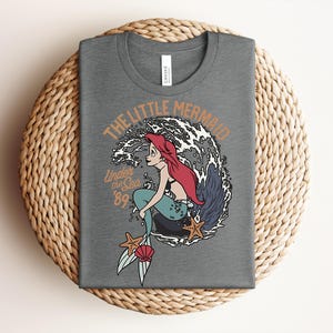 May include: A heather gray t-shirt featuring a graphic of a red-haired mermaid with a teal tail sitting on a rock in front of a large wave. The text reads THE LITTLE MERMAID and Under the Sea '89, accompanied by two orange starfish and a pink seashell.