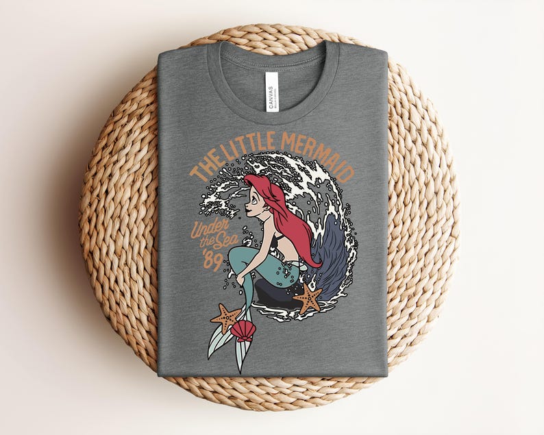 May include: A heather gray t-shirt featuring a graphic of a red-haired mermaid with a teal tail sitting on a rock in front of a large wave. The text reads THE LITTLE MERMAID and Under the Sea '89, accompanied by two orange starfish and a pink seashell.
