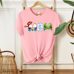 May include: A light pink t-shirt with the word "MOM" in colorful, cartoon-style letters. The "M" features a character with a red hat and yellow flowers. The "O" has a blue sky and cloud design, and the "M" has a green mountain design.