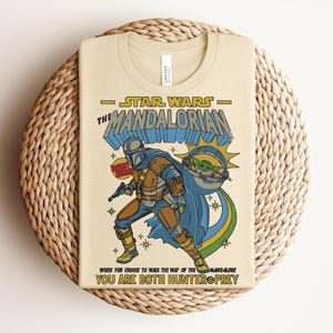 Puede incluir: Una camiseta de color crema con un gr&aacute;fico de The Mandalorian y Grogu. El dise&ntilde;o incluye el texto "Star Wars The Mandalorian", "Bounty Hunter" y "When you choose to walk the way of the Mandalore you are both hunter & prey."