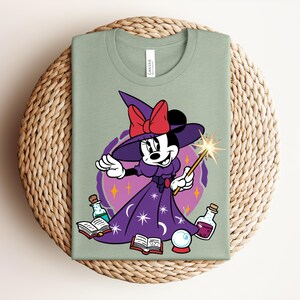 May include: Sage green t-shirt featuring a cartoon character dressed as a witch. The character wears a purple dress, a witch hat with a red bow, and holds a wand. The design includes potion bottles, books, and a crystal ball.