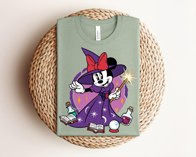 May include: Sage green t-shirt featuring a cartoon character dressed as a witch. The character wears a purple dress, a witch hat with a red bow, and holds a wand. The design includes potion bottles, books, and a crystal ball.