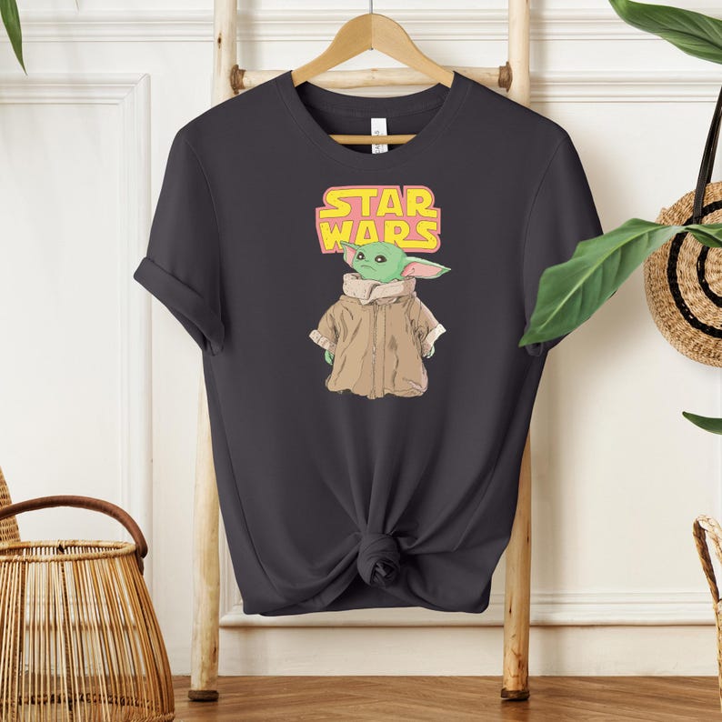 Puede incluir: Camiseta gris oscuro con un gr&aacute;fico de Grogu de Star Wars. El dise&ntilde;o incluye el texto "STAR WARS" en amarillo y rosa sobre un personaje verde de Grogu con un abrigo marr&oacute;n. La camiseta est&aacute; colgada de una percha de madera.