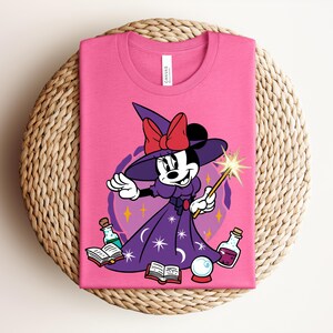 May include: A pink t-shirt featuring a cartoon Minnie Mouse dressed as a witch. She wears a purple dress, witch hat with a red bow, and holds a wand. The design includes potion bottles, books, and a crystal ball.
