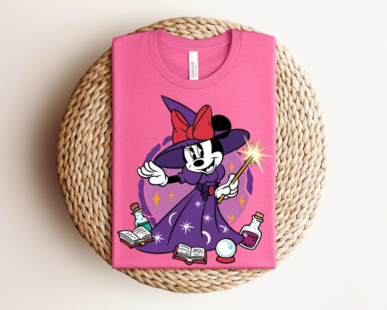 May include: A pink t-shirt featuring a cartoon Minnie Mouse dressed as a witch. She wears a purple dress, witch hat with a red bow, and holds a wand. The design includes potion bottles, books, and a crystal ball.