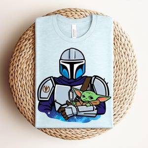 Puede incluir: Camiseta azul claro con una ilustraci&oacute;n de dibujos animados de un personaje mandaloriano sosteniendo a un alien&iacute;gena verde. El dise&ntilde;o incluye un casco, armadura y un fondo espacial. La camiseta est&aacute; doblada sobre una superficie tejida.