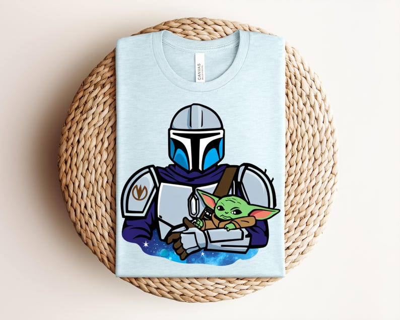 Puede incluir: Camiseta azul claro con una ilustraci&oacute;n de dibujos animados de un personaje mandaloriano sosteniendo a un alien&iacute;gena verde. El dise&ntilde;o incluye un casco, armadura y un fondo espacial. La camiseta est&aacute; doblada sobre una superficie tejida.
