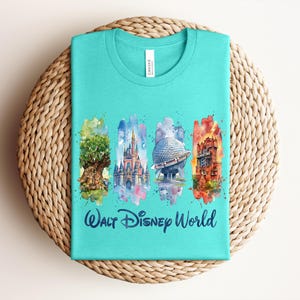 May include: A turquoise t-shirt featuring watercolor illustrations of iconic Walt Disney World landmarks. The design includes the Tree of Life, Cinderella Castle, Spaceship Earth, and Tower of Terror, with "Walt Disney World" text below.