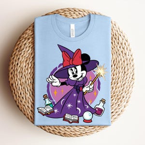 May include: Light blue t-shirt featuring a cartoon Minnie Mouse dressed as a witch, wearing a purple dress and hat with a red bow. She holds a wand and is surrounded by potion bottles, a crystal ball, and books.