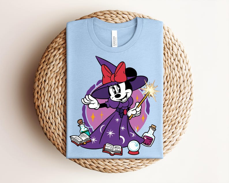 May include: Light blue t-shirt featuring a cartoon Minnie Mouse dressed as a witch, wearing a purple dress and hat with a red bow. She holds a wand and is surrounded by potion bottles, a crystal ball, and books.
