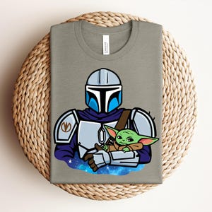 Puede incluir: Una camiseta gris con una ilustraci&oacute;n de dibujos animados de un personaje mandaloriano sosteniendo a un alien&iacute;gena verde. El mandaloriano lleva una armadura plateada con detalles azules. El alien&iacute;gena est&aacute; envuelto en una manta marr&oacute;n, sobre un fondo estrellado.
