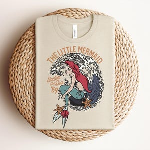 May include: A beige t-shirt featuring a graphic of a red-haired mermaid sitting on a rock in front of a large wave. The text reads THE LITTLE MERMAID, Under the Sea, and 89. Two orange starfish and a pink seashell are positioned near the mermaid's tail.