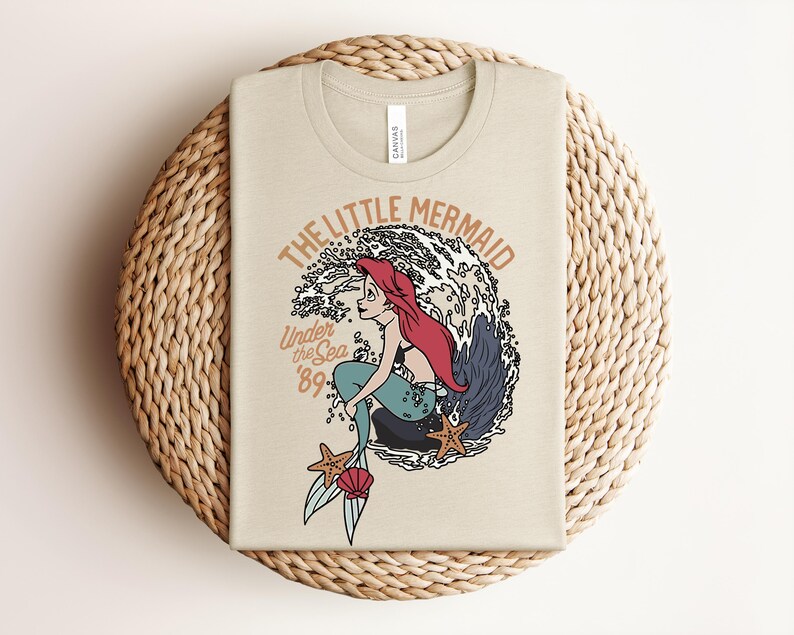 May include: A beige t-shirt featuring a graphic of a red-haired mermaid sitting on a rock in front of a large wave. The text reads THE LITTLE MERMAID, Under the Sea, and 89. Two orange starfish and a pink seashell are positioned near the mermaid's tail.