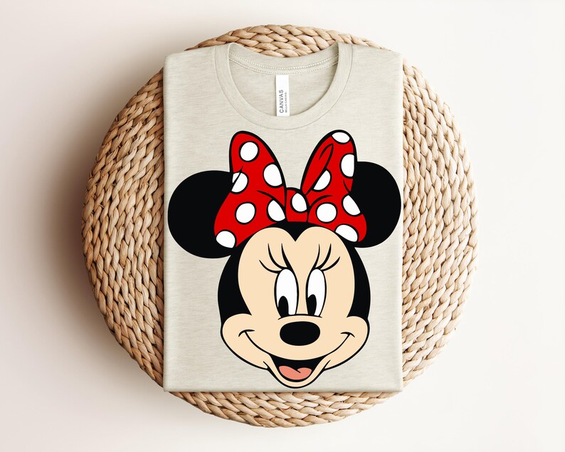 May include: A light beige t-shirt featuring a large graphic of Minnie Mouse's face. The design includes her signature black ears, a red bow with white polka dots, and a smiling expression. The shirt is folded on a woven surface.
