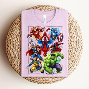 May include: A lilac t-shirt displaying a vibrant graphic of Marvel superheroes such as Iron Man, Captain America, Spider-Man, Thor, Wolverine, Hulk, and Deadpool. The design is framed with a watercolour effect.