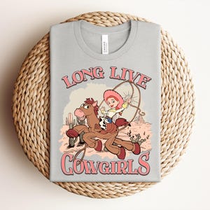 May include: A light gray t-shirt featuring a graphic of a cartoon character riding a brown horse in a desert landscape. The character wears a pink hat and holds a lasso. The text "LONG LIVE COWGIRLS" is printed in pink with a dark outline above and below.