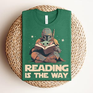 May include: A forest green t-shirt featuring a graphic of a helmeted armored figure sitting cross-legged and reading a book with a small green creature. The text below the graphic reads READING IS THE WAY in large, bold, light-colored capital letters.