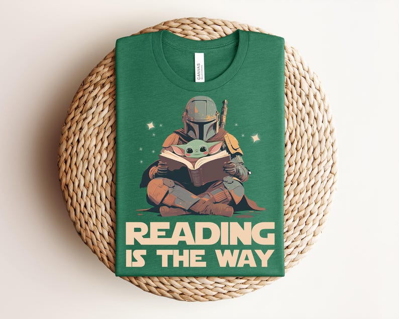 May include: A forest green t-shirt featuring a graphic of a helmeted armored figure sitting cross-legged and reading a book with a small green creature. The text below the graphic reads READING IS THE WAY in large, bold, light-colored capital letters.